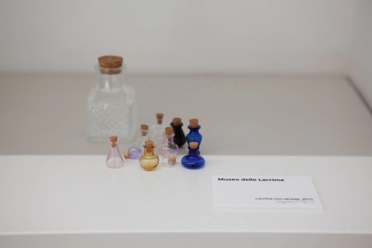 Primo piano di boccette di vetro colorato con tappi di sughero su uno scaffale bianco, con cartellino museale 'Lacrime non versate, 2015' — dettaglio dell'archivio