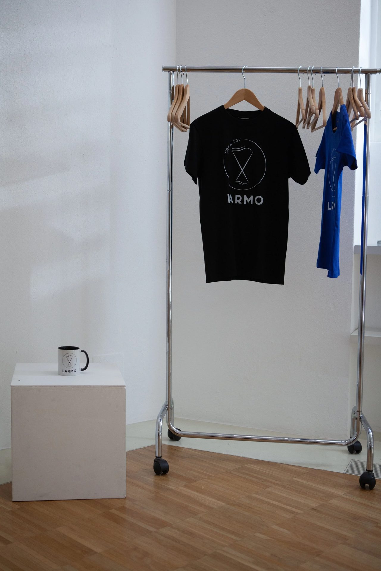 Appendiabiti con t-shirt nera e maglietta blu con logo LARMO, e tazza brandizzata su plinto bianco — vista del merchandise nell'allestimento